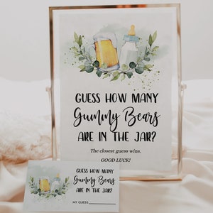 May include: A white sign with black text that reads "Guess how many Gummy Bears are in the jar? The closest guess wins. Good luck!"  The sign is framed in a gold frame and has a watercolour illustration of a beer bottle and a baby bottle with green leaves.