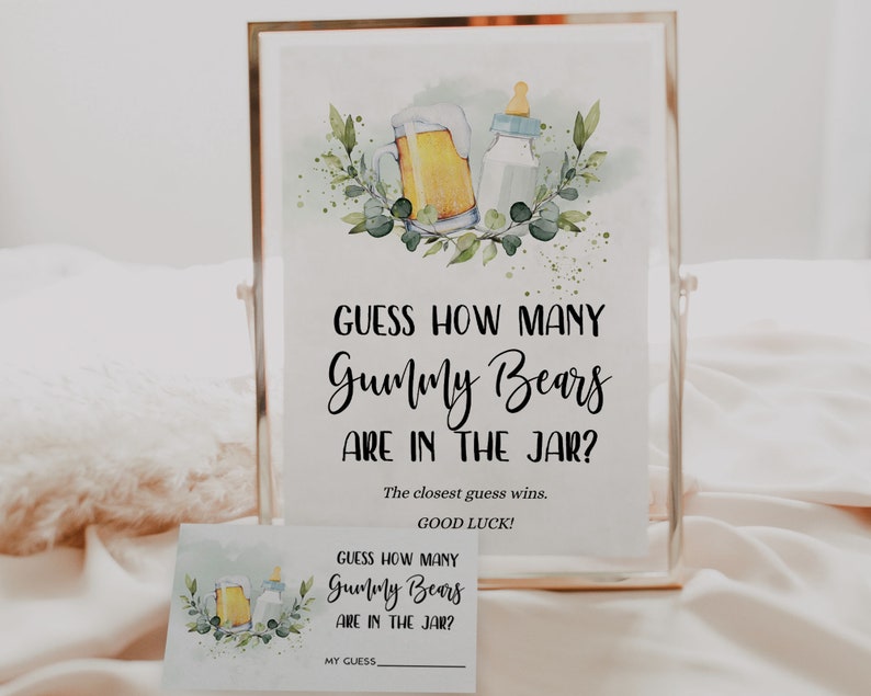 May include: A white sign with black text that reads "Guess how many Gummy Bears are in the jar? The closest guess wins. Good luck!"  The sign is framed in a gold frame and has a watercolour illustration of a beer bottle and a baby bottle with green leaves.