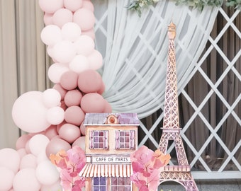 Puede incluir: Una escena decorativa de temática parisina con una Torre Eiffel y un edificio con la etiqueta "CAFE DE PARIS" en estilo acuarela. El fondo incluye un arco de globos rosas, celosía blanca y decoraciones florales.