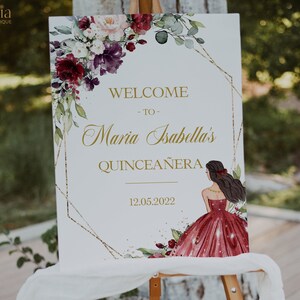 May include: A white welcome sign with gold lettering and a floral design. The sign reads "Welcome to Maria Isabella's Quincea&ntilde;era 12.05.2022".