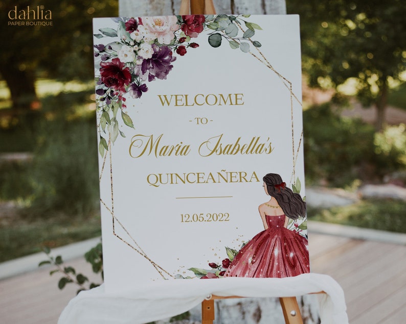 May include: A white welcome sign with gold lettering and a floral design. The sign reads "Welcome to Maria Isabella's Quincea&ntilde;era 12.05.2022".