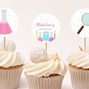May include: Three cupcakes decorated with colorful science-themed cupcake toppers. The toppers feature a beaker, a magnifying glass, and a beaker with the text "Madeline's 8th Birthday".