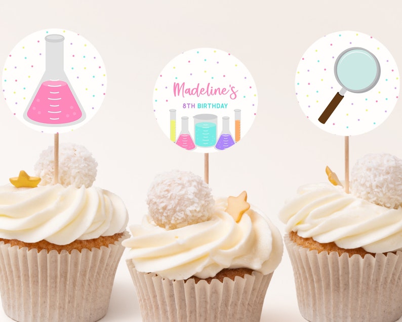 May include: Three cupcakes decorated with colorful science-themed cupcake toppers. The toppers feature a beaker, a magnifying glass, and a beaker with the text "Madeline's 8th Birthday".