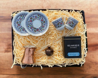 May include: A gift box filled with Turkish coffee supplies. The box contains two small blue and white patterned cups and saucers, a copper coffee pot, a bag of Turkish coffee, and a small decorative pomegranate.
