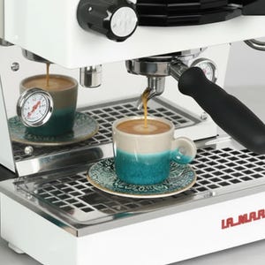 May include: A white espresso machine with a black base and accents. Two small cups are filled with coffee, and a third cup and saucer are on the counter. The machine has a pressure gauge and the brand name "LA MARZOCCO" is visible.