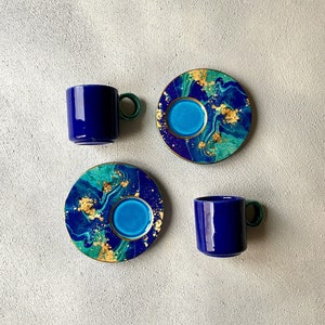 May include: Two deep blue ceramic mugs with green handles paired with two matching saucers. The saucers feature a marbled pattern of blue, teal, and green with gold leaf accents. The mugs have a solid blue exterior and a green handle.
