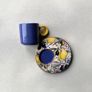 Puede incluir: Una taza de café de cerámica azul con asa amarilla y un platillo a juego con un diseño colorido de limón y aceituna.