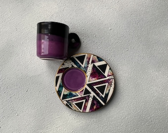 May include: Purple and black ceramic coffee cup with a matching saucer. The saucer has a geometric pattern with black triangles and white dots on a purple background.