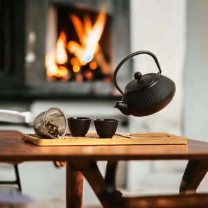May include: A black cast iron teapot with a lid and a spout, pouring tea into three small black teacups. The teapot is sitting on a wooden tray with a tea strainer.