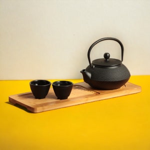Puede incluir: Una tetera de hierro fundido negro con dos tazas de té negras en una bandeja de madera. La tetera tiene una superficie texturizada y un asa redonda. Las tazas de té son pequeñas y redondas. La bandeja está hecha de bambú y tiene un acabado de madera natural.
