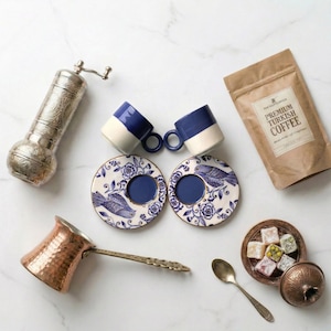 Könnte beinhalten: Eine Auswahl an türkischen Kaffeeartikeln: blaue und weiße Tassen, passende Untertassen, eine Kupferkaffeekanne, eine Kaffeemühle, eine Kaffeetüte und türkisches Lokum. Die Untertassen haben ein blaues Vogelmuster.