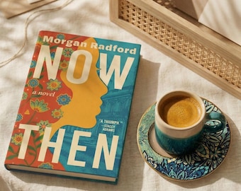 May include: A hardcover book titled Now Then by Morgan Radford with a colorful cover featuring a yellow silhouette, floral patterns, and blue architectural details, placed next to a ceramic mug filled with coffee on a matching patterned saucer.