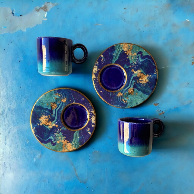 Puede incluir: Tres tazas de caf&eacute; y platillos azules y dorados. Las tazas y los platillos tienen un dise&ntilde;o marmoleado con remolinos azules, dorados y blancos.