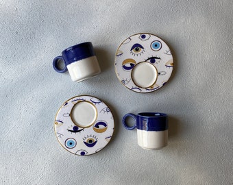 May include: Two white ceramic coffee cups with blue rims and a blue and white eye pattern. The cups are on matching saucers with the same eye pattern.