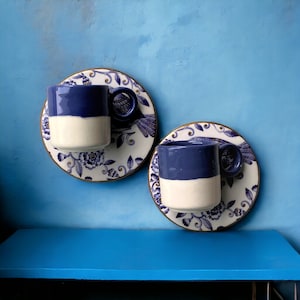 Puede incluir: Dos tazas de café de cerámica azul y blanca con diseños florales en los platillos. Las tazas tienen una banda blanca alrededor del medio y una parte superior azul.