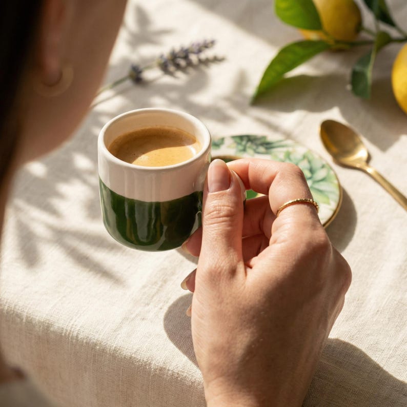 Close up of a woman's hand holding a small green and white ceramic espresso cup, bringing it to her lips to take a sip in the morning sunlight.