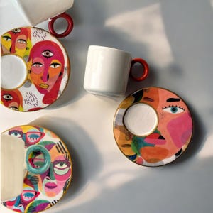 May include: Set of three colorful ceramic coffee cups and saucers with abstract face designs. Each saucer features a different face with bold colors and patterns. The cups are white with red handles.