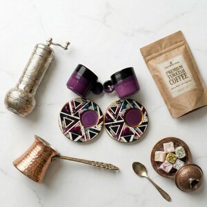 May include: A collection of Turkish coffee items, including two purple and black espresso cups, matching saucers, a copper coffee pot, a silver grinder, a bag of coffee, and a small dish of Turkish delight.