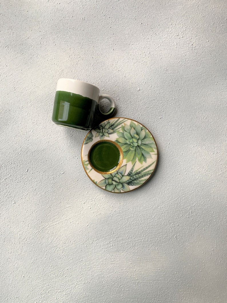 Pu&ograve; includere: Un set di tazza e piattino in ceramica. La tazza &egrave; verde scuro con un bordo bianco, e il piattino presenta un design di piante succulente con finiture dorate. Il set &egrave; su una superficie bianca strutturata.
