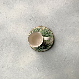 Pu&ograve; includere: Una tazza da t&egrave; in ceramica bianca poggia su un piattino abbinato con un design di piante succulente. Il piattino ha un bordo dorato e la tazza &egrave; vuota. Lo sfondo &egrave; una superficie strutturata azzurra.