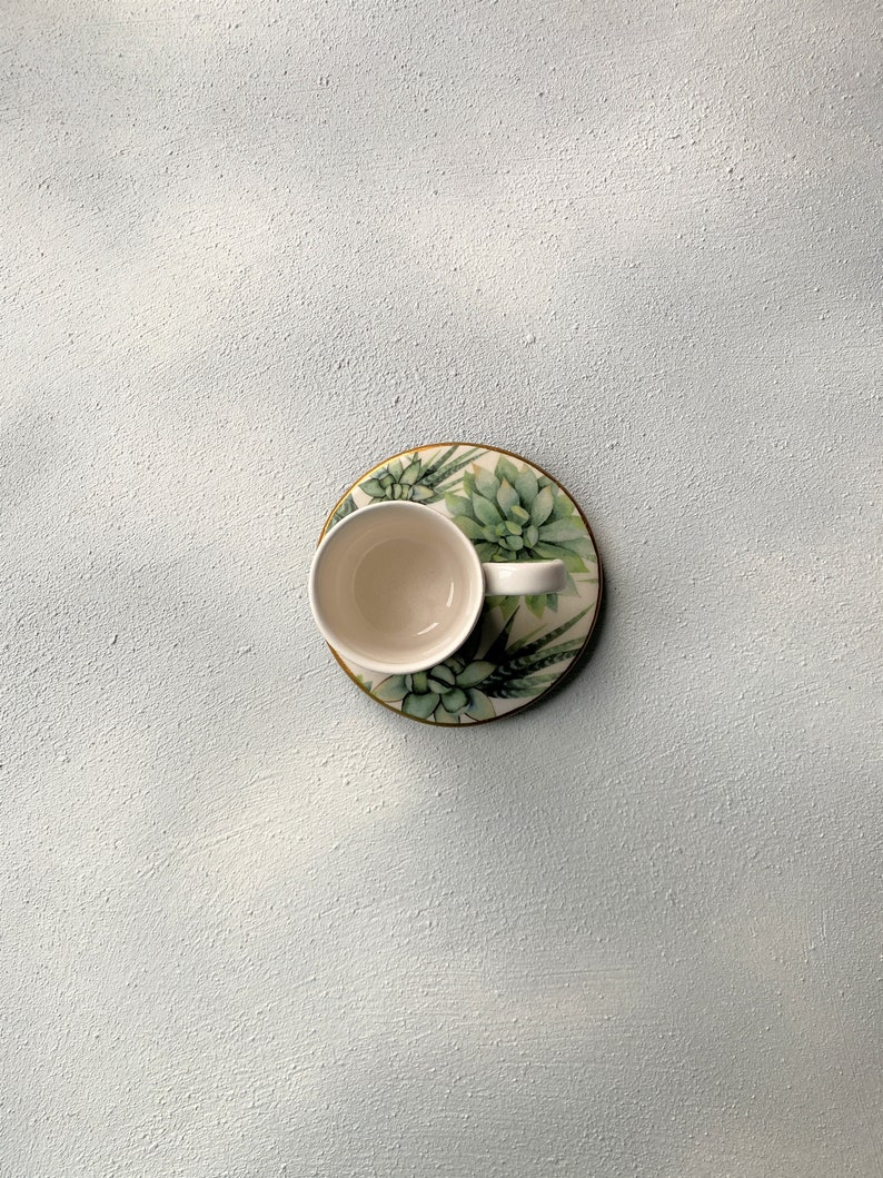 Pu&ograve; includere: Una tazza da t&egrave; in ceramica bianca poggia su un piattino abbinato con un design di piante succulente. Il piattino ha un bordo dorato e la tazza &egrave; vuota. Lo sfondo &egrave; una superficie strutturata azzurra.