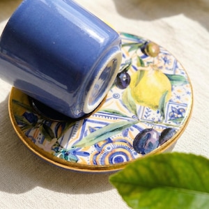 Extreme close up of a glossy ceramic espresso cup showing hand-painted blue and yellow lemon details and a shiny gold rim in bright sunlight.