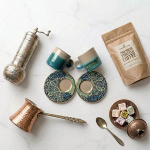 May include: An assortment of Turkish coffee items, including two small blue and beige mugs with matching saucers, a copper coffee pot, a silver coffee grinder, a bag of coffee, and a small dish of Turkish delight.