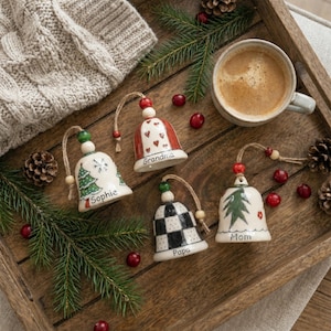 May include: Four ceramic bell ornaments with twine hangers, each personalized with names including Sophie, Grandma, Papa, and Mom. The bells feature hand-painted designs such as a green Christmas tree, red hearts, and a black and white checkerboard pattern.
