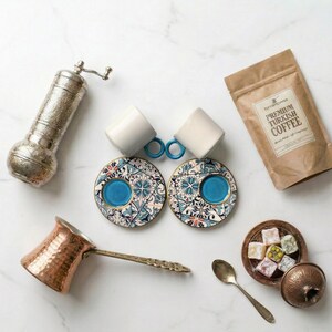May include: An overhead shot of a Turkish coffee set. The set includes two white coffee cups with blue handles, two patterned saucers, a copper coffee pot, a silver coffee grinder, a bag of coffee, and a small dish of Turkish delight.
