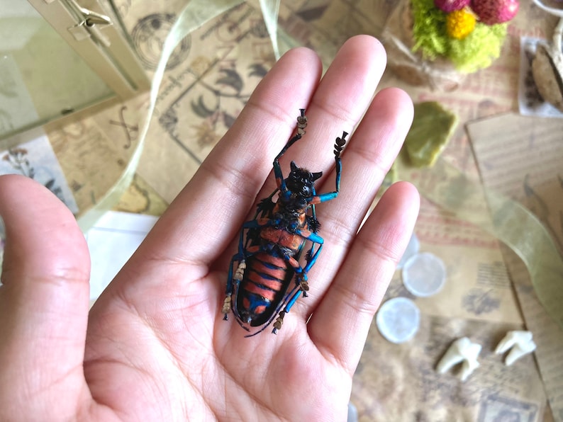 May include: A detailed, colorful insect model with blue, orange, and black accents is held in an open hand. The insect's intricate design is visible, showcasing its legs and body structure. The background includes decorative elements.