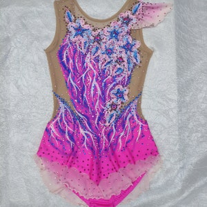 May include: A sleeveless rhythmic gymnastics leotard in shades of pink and purple, adorned with floral appliqu&eacute;s and sparkling embellishments. The bodice features a nude mesh overlay, and the skirt has a layered, scalloped hem. The design includes blue and pink crystals.