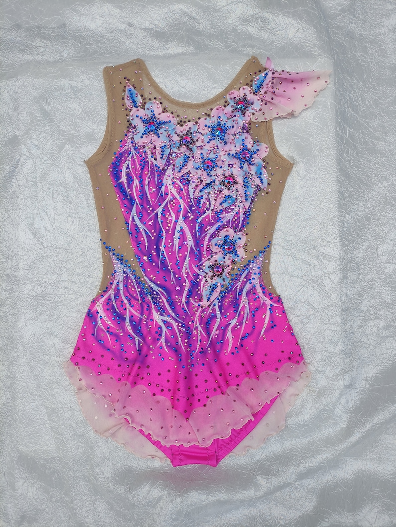 May include: A sleeveless rhythmic gymnastics leotard in shades of pink and purple, adorned with floral appliqu&eacute;s and sparkling embellishments. The bodice features a nude mesh overlay, and the skirt has a layered, scalloped hem. The design includes blue and pink crystals.
