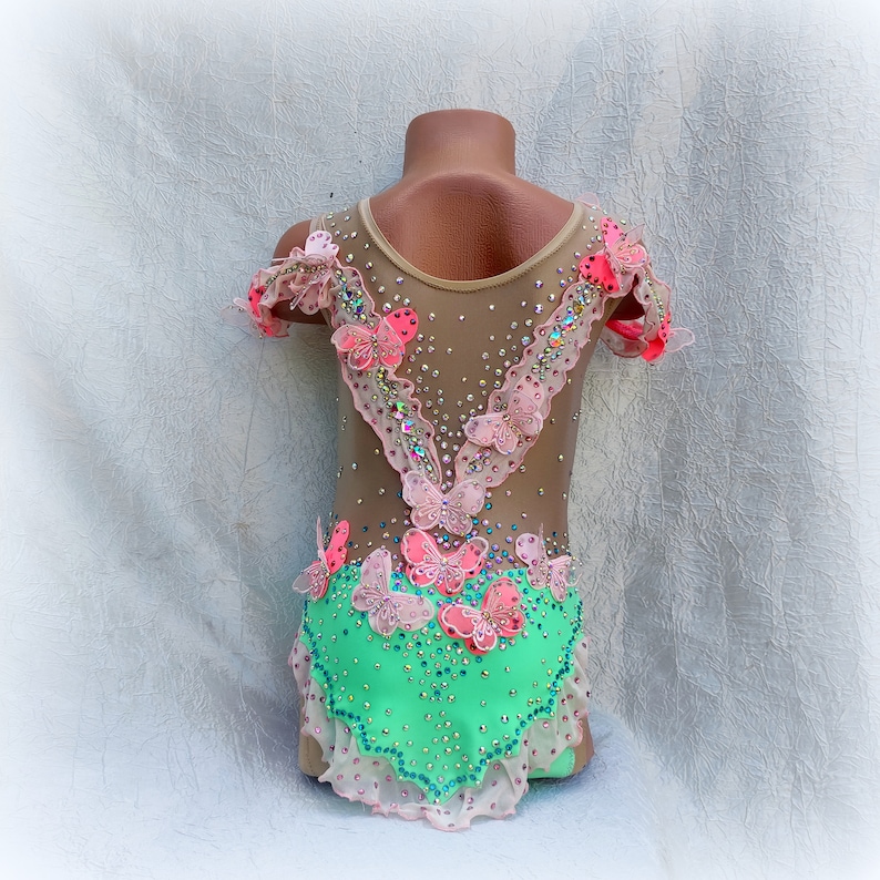 May include: A light green leotard with pink and light pink butterfly embellishments. The leotard has a sheer mesh bodice with a V-neckline and short sleeves.