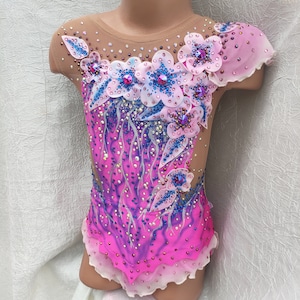 Puede incluir: Maillot rosa y morado con diseño floral. El maillot presenta un escote y mangas transparentes, con un efecto degradado de rosa a morado. El diseño floral incluye flores blancas y azules con adornos de cristal.