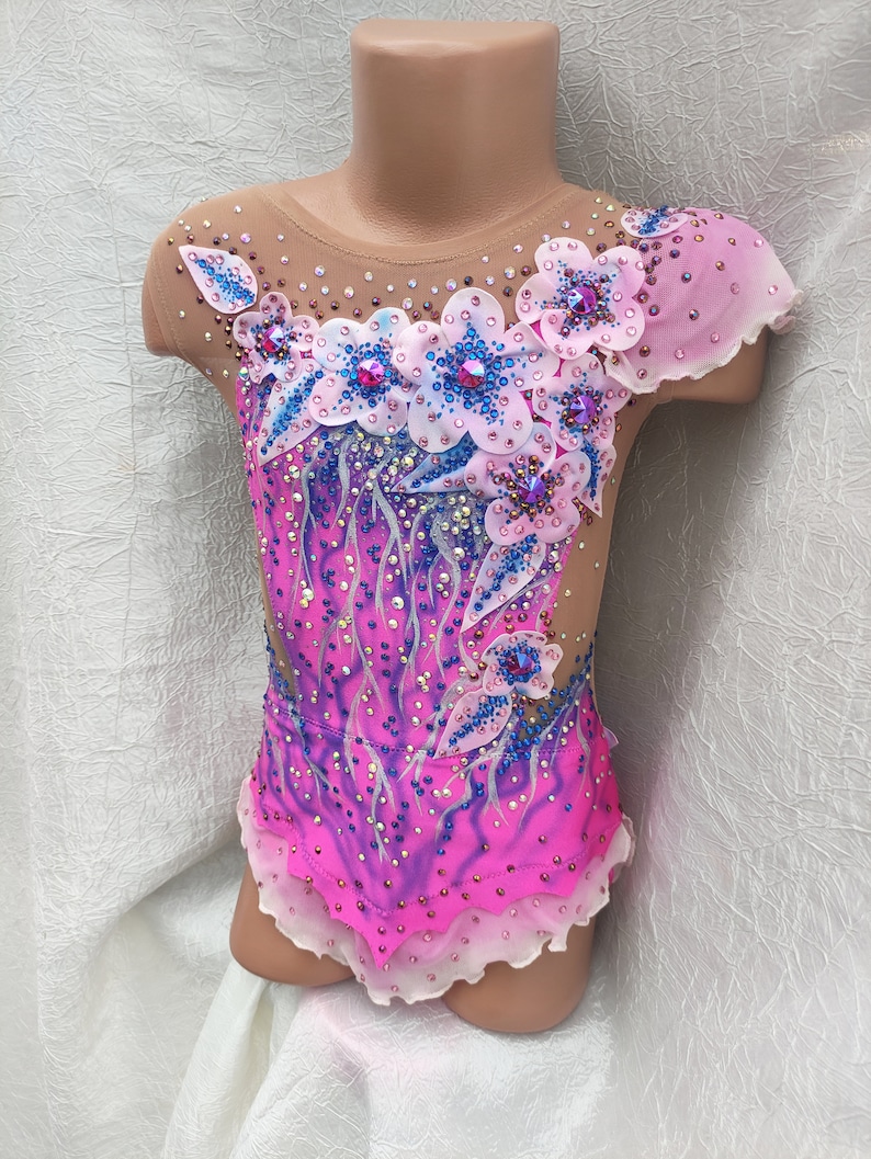 May include: A pink and purple leotard with a floral design. The leotard features a sheer neckline and sleeves, with a gradient effect from pink to purple. The floral design includes white and blue flowers with crystal embellishments.
