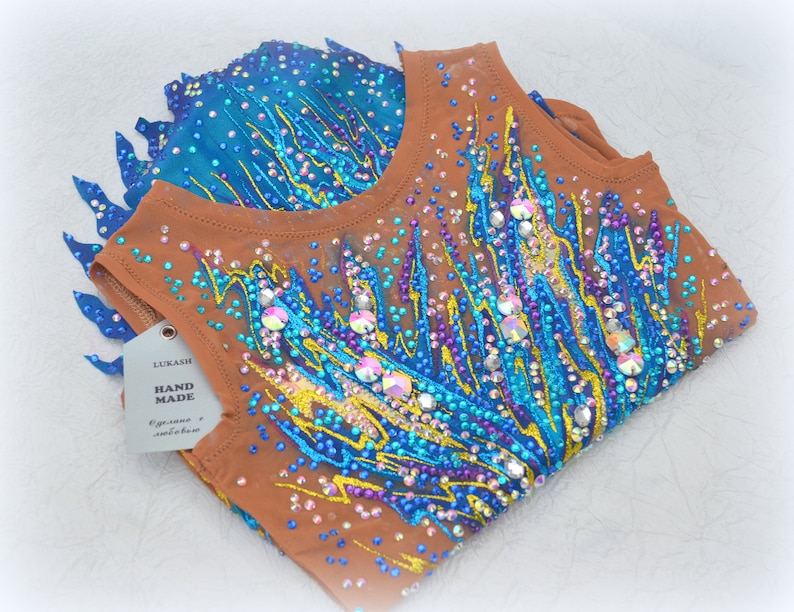 May include: A brown leotard with a blue, yellow, and purple flame design. The leotard is covered in rhinestones and has a tag that says "LUKASH HAND MADE".