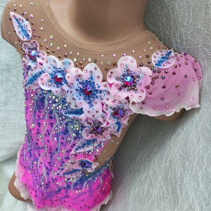 May include: A pink and purple leotard with floral embellishments. The leotard features a gradient design, transitioning from a vibrant pink to a deep purple. Delicate white flowers with blue accents and sparkling rhinestones adorn the bodice and sleeves.