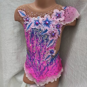 May include: A vibrant pink and purple leotard with floral embellishments. The bodice features a gradient design and is adorned with sparkling rhinestones and delicate flower appliqu&eacute;s in shades of pink, blue, and white. The sleeves are sheer.