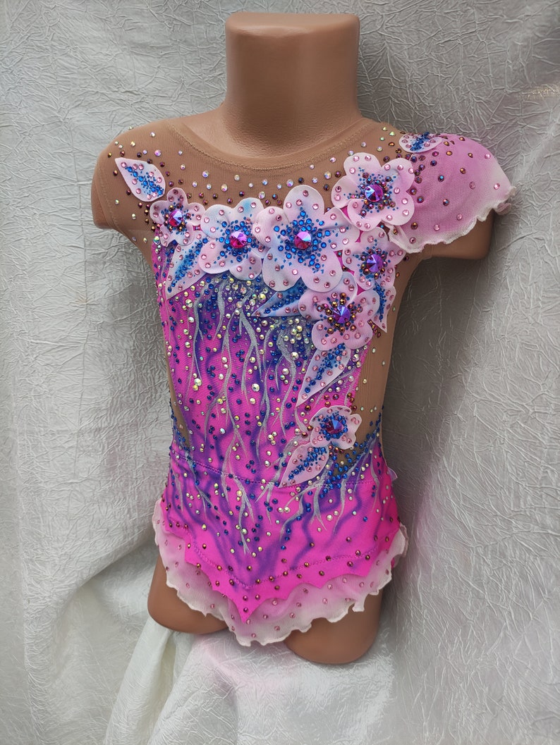 May include: A vibrant pink and purple leotard with floral embellishments. The bodice features a gradient design and is adorned with sparkling rhinestones and delicate flower appliqu&eacute;s in shades of pink, blue, and white. The sleeves are sheer.