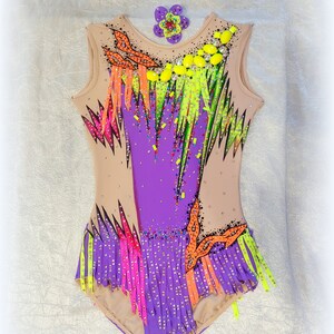 May include: A purple and beige leotard with colorful fringe and rhinestones. The leotard has a sleeveless design and a scoop neck. A small flower embellishment is attached to the front of the leotard.