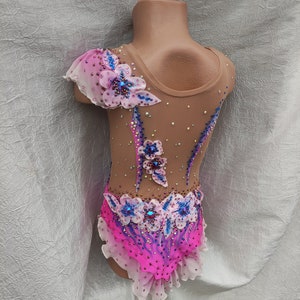 May include: A tan leotard with pink and blue floral embellishments. The leotard features a high neckline and is adorned with rhinestones and a ruffled skirt. The design includes a sheer, one-shoulder sleeve with a floral accent.