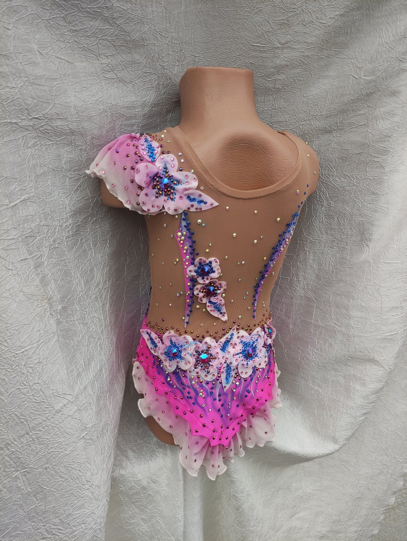 May include: A tan leotard with pink and blue floral embellishments. The leotard features a high neckline and is adorned with rhinestones and a ruffled skirt. The design includes a sheer, one-shoulder sleeve with a floral accent.