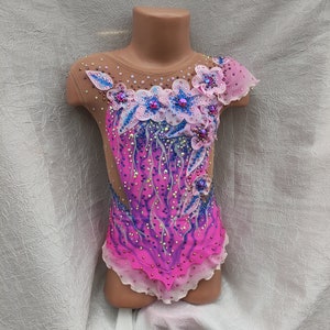 May include: A vibrant pink and purple leotard for rhythmic gymnastics, adorned with floral appliqu&eacute;s, rhinestones, and a sheer mesh neckline and sleeves. The design features a gradient effect and delicate embellishments.