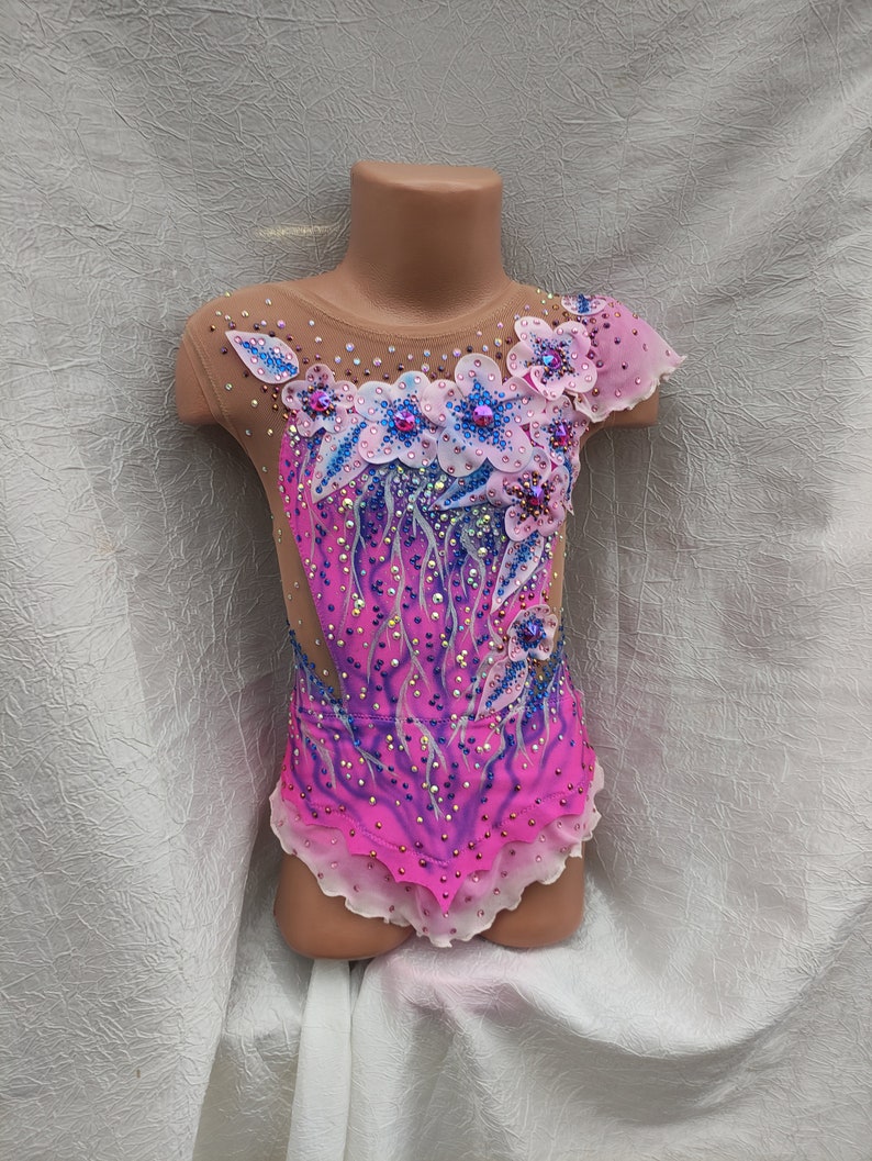 May include: A vibrant pink and purple leotard for rhythmic gymnastics, adorned with floral appliqu&eacute;s, rhinestones, and a sheer mesh neckline and sleeves. The design features a gradient effect and delicate embellishments.