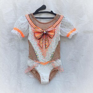 May include: A white and nude leotard featuring short puff sleeves with orange trim, a large orange and white patterned bow at the centre chest, and extensive crystal embellishments throughout the bodice and sleeves on a black hanger.