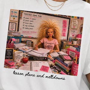 May include: A white t-shirt featuring a graphic print of a doll with wild blonde hair sitting at a desk covered in school supplies, papers, and a mug. The text on the graphic reads Teacher Game Plan and Running on Caffeine, with the phrase lesson plans and meltdowns printed below.