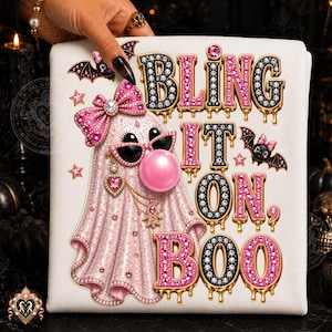 May include: A white t-shirt featuring a graphic of a ghost wearing a pink bow, heart-shaped earrings, and sunglasses while blowing a pink bubble. The text reads BLING IT ON, BOO in black and pink jewelled letters with gold drips and small bats.