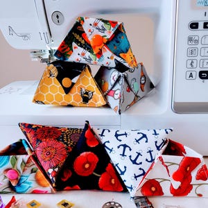 May include: Handmade fabric triangle pouches in diverse patterns, including floral, cat, bee, and anchor designs. Each pouch has a button closure. The pouches are displayed on a white surface near a sewing machine.