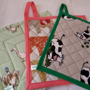 May include: Three quilted pot holders with different designs. The first is green with a white duck pattern, the second is pink with a brown and white animal pattern, and the third is beige with a black and white cow pattern.