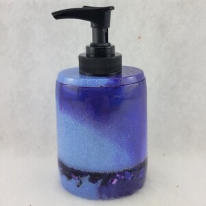 Può includere: Un dispenser di sapone viola e blu con glitter. Il dispenser ha una pompa nera e una base trasparente.